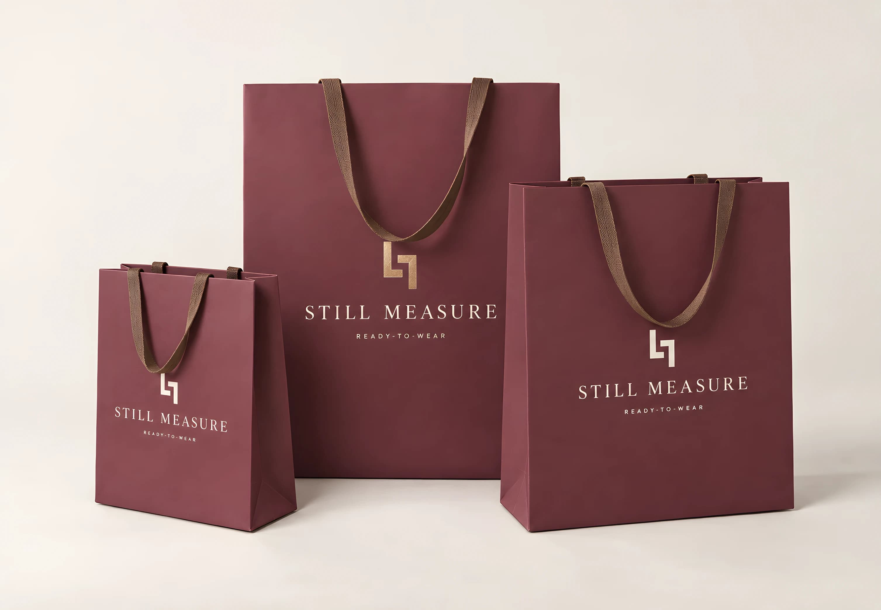 Three mulberry-wine paper shopping bags of three sizes on an oyster studio sweep, with flat tobacco-brown ribbon handles. Each front panel carries the two-L mark above STILL MEASURE and READY-TO-WEAR — the mark in champagne gold foil on the tallest bag and in oyster white on the two smaller ones.