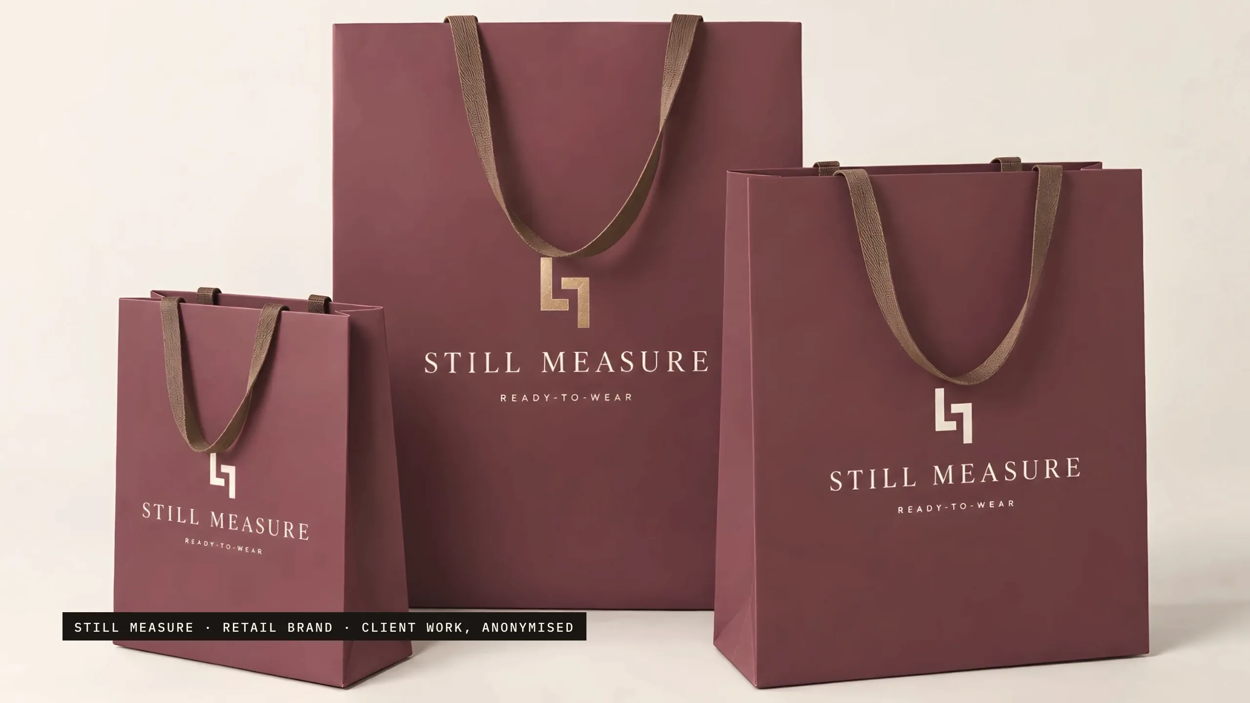 Three mulberry-wine paper shopping bags of three sizes on an oyster studio sweep, with tobacco-brown ribbon handles, each carrying the two-L mark above STILL MEASURE and READY-TO-WEAR.